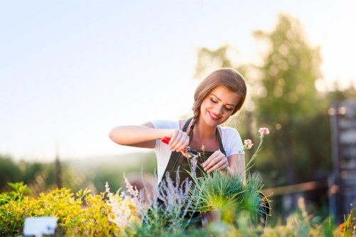 A woman with light brown hair and a smile is tending to a garden bed, using a small garden fork to work among various plants and flowers. She is dressed in a light blue top and white apron, standing outdoors during daylight hours with soft sunlight illuminating the scene. The garden features a mix of flowering plants, small shrubs, and patches of grass with well-defined flower beds bordered by soil and mulch. In the background, there are blurred trees and a blue sky, indicating a peaceful outdoor environment typical of a private or community garden in Muswell Hill. The plants display a variety of natural green tones, complemented by colorful flowers, and the garden appears well-maintained, reflecting professional gardening care services. The overall setting emphasizes a well-kept, landscaped outdoor space suitable for lawn and garden upkeep, landscaped with attention to detail typical of local gardening service standards.