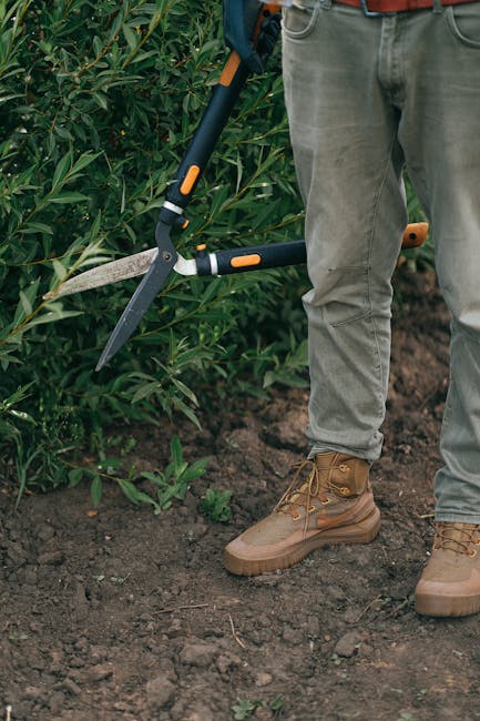 A gardener from gardeningservicesmuswellhill.co.uk is working in a neatly maintained garden, standing on dark, moist soil with a slightly uneven texture. The gardener's lower body is visible, dressed